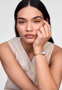 Reloj de metal dorado con correa trenzada, esfera blanca y marcadores de hora sencillos. El modelo lleva anillos adicionales de oro y plata. Atuendo de tonos neutros.