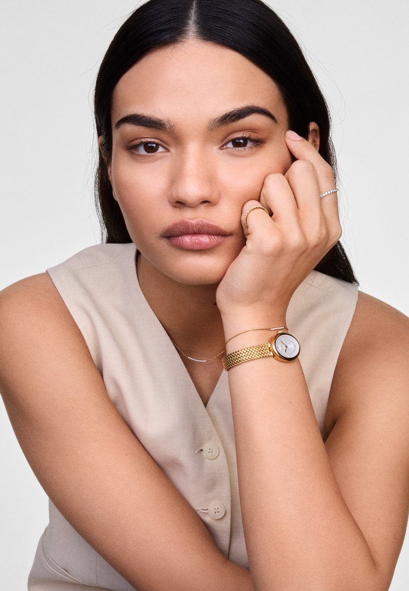 Reloj de metal dorado con correa trenzada, esfera blanca y marcadores de hora sencillos. El modelo lleva anillos adicionales de oro y plata. Atuendo de tonos neutros.