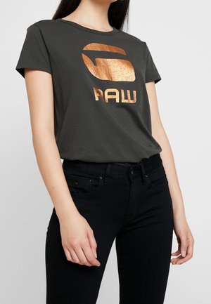 Frau trägt dunkelgraues T-Shirt mit glänzendem kupferfarbenem "RAW"-Logo und schwarze Jeans, steht vor einfachem Hintergrund.
