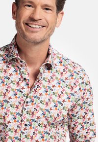 Chemise à manches courtes à motifs floraux avec un fond blanc ornée de motifs colorés de fleurs en rose, orange, bleu et vert, avec des boutons noirs.