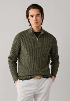 Jonge man met donker haar die een olijfgroene geribbelde trui met rits draagt en lichtbeige broek met handen in de zakken.