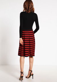 Femme aux cheveux mi-longs portant un haut noir à manches longues, une jupe rouge et noire à rayures horizontales arrivant au genou, et des sandales à talons hauts noires.