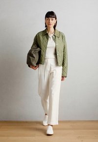 Veste matelassée verte avec des pressions, portée sur un top blanc texturé et un pantalon large blanc, assortie de baskets blanches.