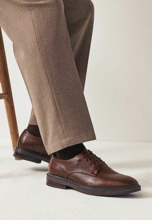 Chaussures habillées en cuir marron portées avec des chaussettes noires et un pantalon beige, montrées avec un pied posé sur le pied en bois d'une chaise.
