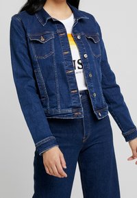 Jeansjacka med en mörkblå tvätt, klassisk krage, knäppning med knappar, bröstfickor och kontrasterande sömmar på ärmar och nederkant.