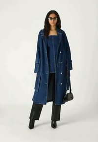 Μακρύ τζιν παλτό με άσπρες ραφές, διαθέτει μεγάλα κουμπιά, πλαινές τσέπες και ανοιχτό μπροστινό μέρος. Φοριέται πάνω από τζιν μπλούζα και μαύρους παντελόνες.