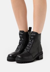 Černé kožené kotníkové boty s robustní podrážkou, vybavené zipovým uzávěrem a ozdobnými popruhy se značkou DKNY. Hladký povrch a pevný design.