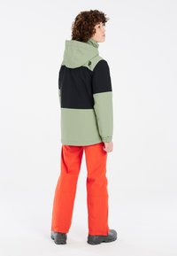 Veste verte et noire avec capuche, présentant un design coloré. Associée à un pantalon orange vif et des chaussures noires.