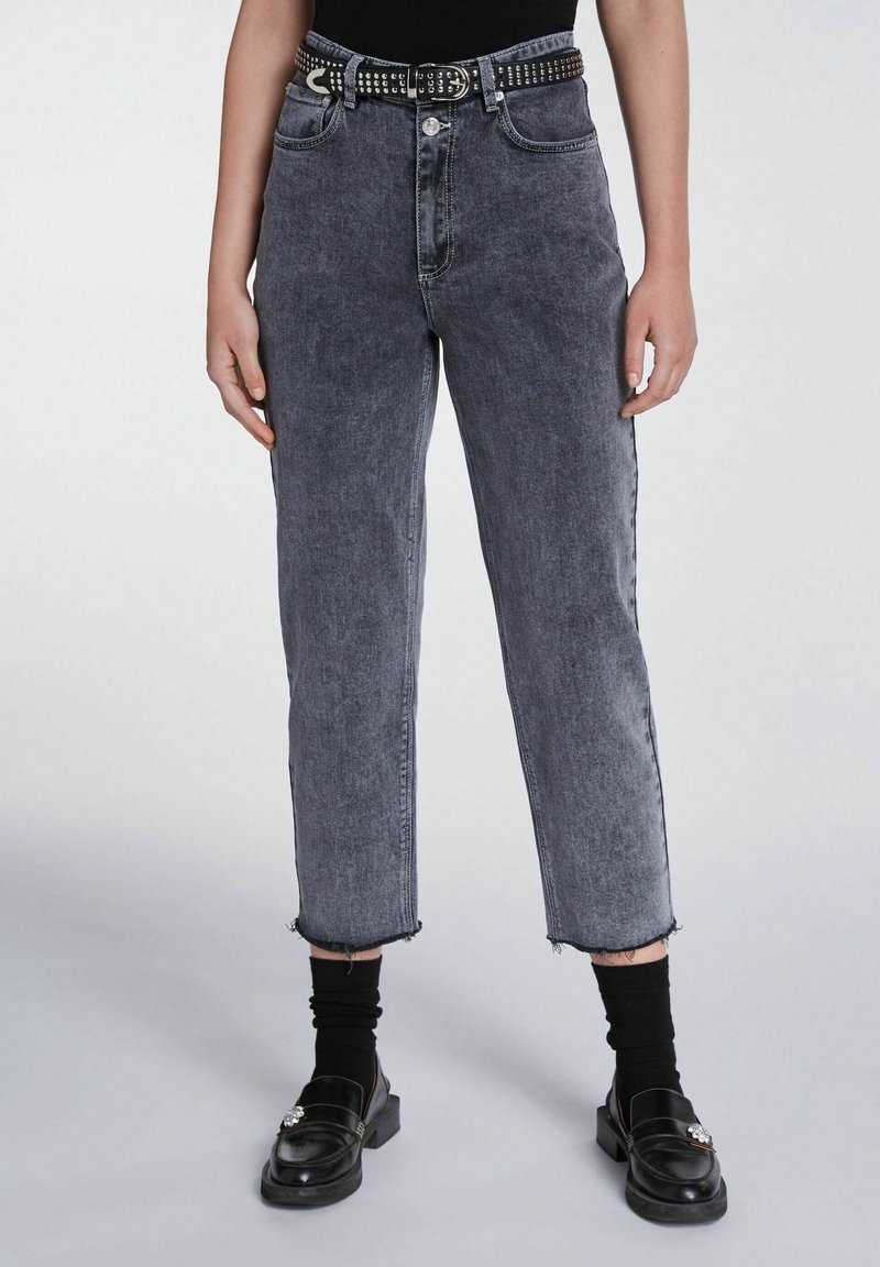 Jeans in denim nero sbiadito a vita alta con orli sfrangiati, abbinati a una cintura nera borchiata, indossati con calzini neri e mocassini.