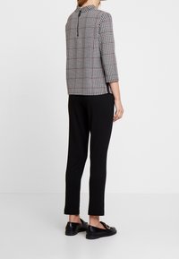 Donna in piedi con indosso un top a scacchi bianco e nero con maniche tre quarti, pantaloni neri slim e mocassini neri con fibbie dorate.