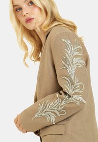 Blazer beige con tejido texturizado que presenta patrones de hojas con lentejuelas en las mangas. Diseño entallado con solapa muestrada y dos bolsillos.