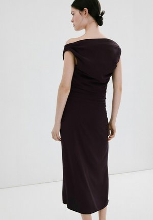 Vrouw met donker haar in een lage knot, gekleed in een nauwsluitende, paarsachtige midi-jurk met blote schouders, staand tegen een effen witte achtergrond.