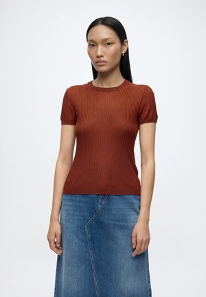 Kvinde med langt sort hår iført små guldøreringe, en figursyet brun ribbet top med korte ærmer og et blåt denimskørt, stående mod en hvid baggrund.