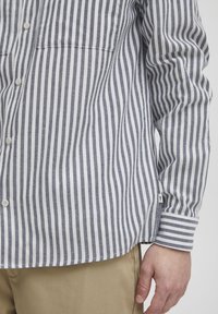 Camicia con bottoni caratterizzata da righe orizzontali blu navy e bianche, maniche lunghe, una tasca sul petto e un orlo che curva leggermente ai lati.