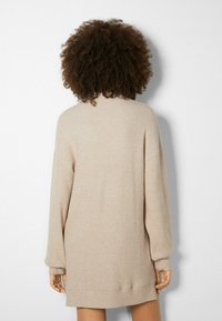 Beige stickad klänning med långa ärmar, ribbad textur och avslappnad passform, som når till mitten av låret. Sömlös design och neutral färg.