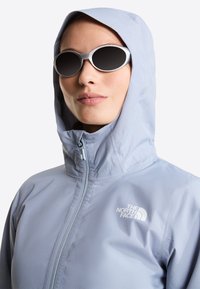 Γυναίκα που φοράει μπουφάν North Face ανοιχτού γαλάζιου χρώματος με κουκούλα, φερμουάρ κλειστό μέχρι το λαιμό, και οβάλ γυαλιά ηλίου με λευκή πλαίσια και σκοτεινούς φακούς.