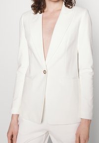Biała marynarka z głębokim dekoltem w kształcie V, z pojedynczym złotym zapięciem na guzik, dwiema przednimi kieszeniami, dopasowanym krojem i gładką fakturą tkaniny.