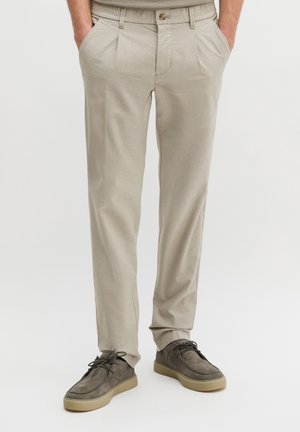 Mann trägt beigefarbene Plisseehose, die Hände in den Taschen, und dunkelgraue Wildleder-Schnürschuhe vor weißem Hintergrund.