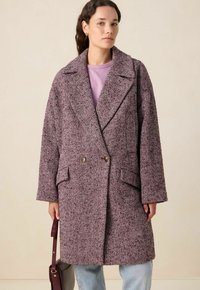 Manteau croisé avec un motif pied-de-poule, doté d'un large col, de poches latérales et d'un tissu bordeaux texturé. Porté par-dessus un haut léger.
