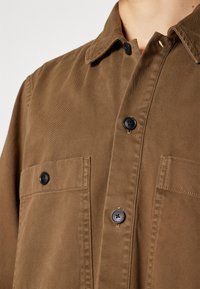 Gros plan d'une chemise marron boutonnée avec des boutons noirs et deux poches poitrine, portée par une personne à la peau claire.
