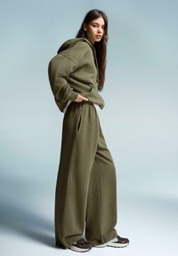 Olivově zelená mikina s oversized střihem a bočními kapsami, v kombinaci s širokými joggingovými kalhotami stejné barvy. Hnědé sneakers s bílými akcenty.
