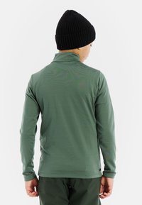 Camicia leggera verde a maniche lunghe con colletto alto, texture morbida e design aderente. Indossata con un berretto a coste nero.