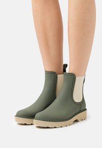 Stivali alla caviglia impermeabili verdi con pannelli elasticizzati beige e suola in tessuto marrone chiaro, dotati di una linguetta sul retro per una facile calzata.