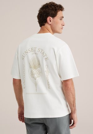 Mann mit kurzen lockigen Haaren, der ein weißes übergroßes T-Shirt mit beiger botanischer Grafik und Text auf dem Rücken trägt, kombiniert mit grauer Hose.