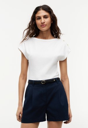 Frau mit welligem braunem Haar, trägt ein weißes kurzärmeliges Oberteil mit goldenen Knöpfen und marineblaue Shorts mit Gürtel, steht vor einem schlichten Hintergrund.