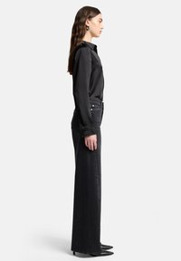 Czarna satynowa koszula z długim rękawem w zestawieniu z ciemnymi dżinsami o wysokim stanie, ozdobionymi perforowanym wzorem po bokach, oraz czarnymi szpiczastymi szpilkami.