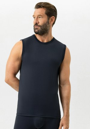 Man met baard die een mouwloos donkerblauw hemd draagt, kijkt opzij en staat tegen een lichte achtergrond.