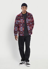 Veste chemise à motifs multicolores en rouge, bleu et noir avec une fermeture à boutons, associée à une chemise noire et un jean foncé. Baskets noires.