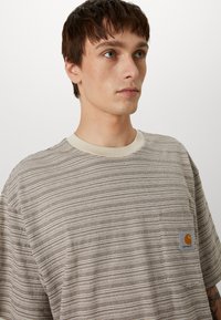 Striped short-sleeve t-shirt in beige and brown tones, featuring a chest pocket with a logo and a crew neck. Cotton material with textured fabric.