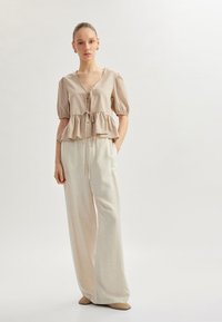 Haut peplum beige clair avec des rayures verticales, manches bouffantes courtes et fermeture à cordon. Associé à un pantalon large léger de couleur claire.