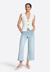Top sin mangas blanco con escote en V, que presenta tres botones negros. Jeans de pierna ancha de un azul claro con un dobladillo sin terminar. Zapatos beige de punta afilada.