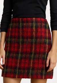 Raudona, juoda ir žalia vilnonė tartano sijonas, pasižymintis pūkuota tekstūra, tiesiu kirpimu ir matomu hemeliu, siekiančiu iki vidurio šlaunų.