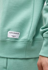 Κοντινό ενός ατόμου που φοράει φούτερ και παντελόνι σε μέντα πράσινο χρώμα με λευκή ετικέτα Björn Borg που αναγράφει «Stockholm 1956» στο φούτερ.