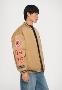 Bežna bomber jakna s črnimi črtami, ki ima rdeče tekstilne nalepke "04" in "25" na rokavu ter podrobnost z logotipom na prsih. Enostavna tekstura.