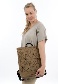 Mujer con cabello rubio y blusa beige sosteniendo una mochila Suri Frey marrón con patrón geométrico sobre un fondo blanco liso.