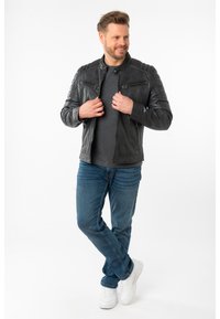 Schwarze Leder-Motorradjacke für Herren mit gesteppten Schulterdetails, zwei vorderen Reißverschlusstaschen und einer taillierten Passform, kombiniert mit blauen Jeans und weißen Sneakers.