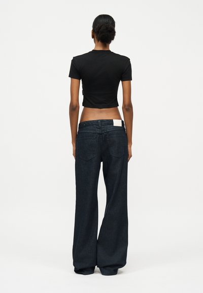 Άτομο με σκούρα μαλλιά δεμένα πίσω, ντυμένο με μαύρο κοντομάνικο τοπ και φαρδιά σκούρα μπλε τζιν, που στέκεται με την πλάτη σε απλό φόντο.