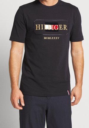 Homme portant un t-shirt noir à manches courtes avec le logo "HILFIGER" et les chiffres romains MCMLXXXV sur la poitrine, associé à un pantalon sombre à rayures fines.