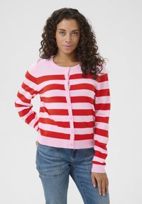 Röd och rosa randig cardigan, rundad hals, knappad fram, ribbade ärmar och nederkant, tillverkad av mjukt stickat material. Bärs över blå jeans.