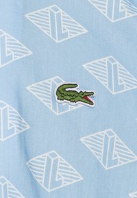 Lichtblauwe stof met een patroon van witte geometrische vormen en een kleine groene geborduurde krokodil met een open rode mond.