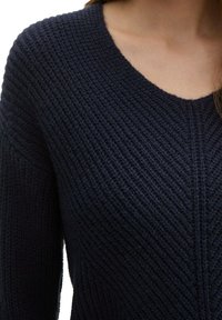 Granatowy sweter robiony na drutach z dekoltem w kształcie litery V, z fakturowanym ribbingiem oraz ukośnymi wzorami wzdłuż rękawów i korpusu.