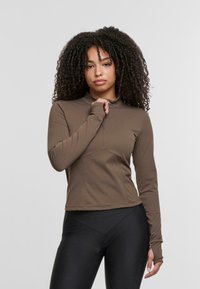 Braun langärmeliges Oberteil mit hohem Kragen und taillierter Passform, versehen mit einem verdeckten Reißverschluss und glattem, elastischem Stoff. Darunter schwarze Leggings.