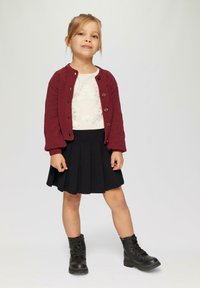 Cardigan bourgogne tricoté avec des boutons, chemise blanche avec des motifs de cœurs, jupe plissée noire et bottes noires à lacets.