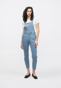 Blå denimdress med justerbare stropper, knappe detaljer og en litt innsnevret benform. Brukt over en lys kremfarget skjorte.