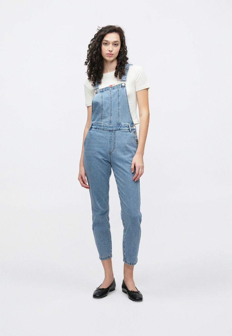 Blå denimdress med justerbare stropper, knappe detaljer og en litt innsnevret benform. Brukt over en lys kremfarget skjorte.