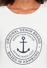 T-shirt branca com um logótipo circular azul-marinho com uma âncora, texto detalhando a marca "Tom Tailor" e o ano de fundação 2007.